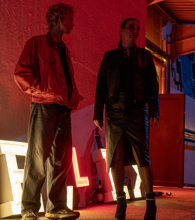 "SOKO Potsdam - Trixi": In einer nächtlichen Szene stehen Trixi Böttcher (Annika Kuhl) und ihr Sohn Janik (Philip Günsch) auf einer Rampe vor einem Club. Neonlicht und rote Strahler tauchen die Betonwände und die Figuren in warmes, intensives Licht. Janik trägt eine weite Jeans und eine rote Jacke, Trixi ein schwarzes Lederkostüm mit engem Rock und hohen Schuhen. Neben ihren Füßen leuchten große, angeschaltete Buchstaben des Clubnamens, Flaschen stehen auf dem Boden.