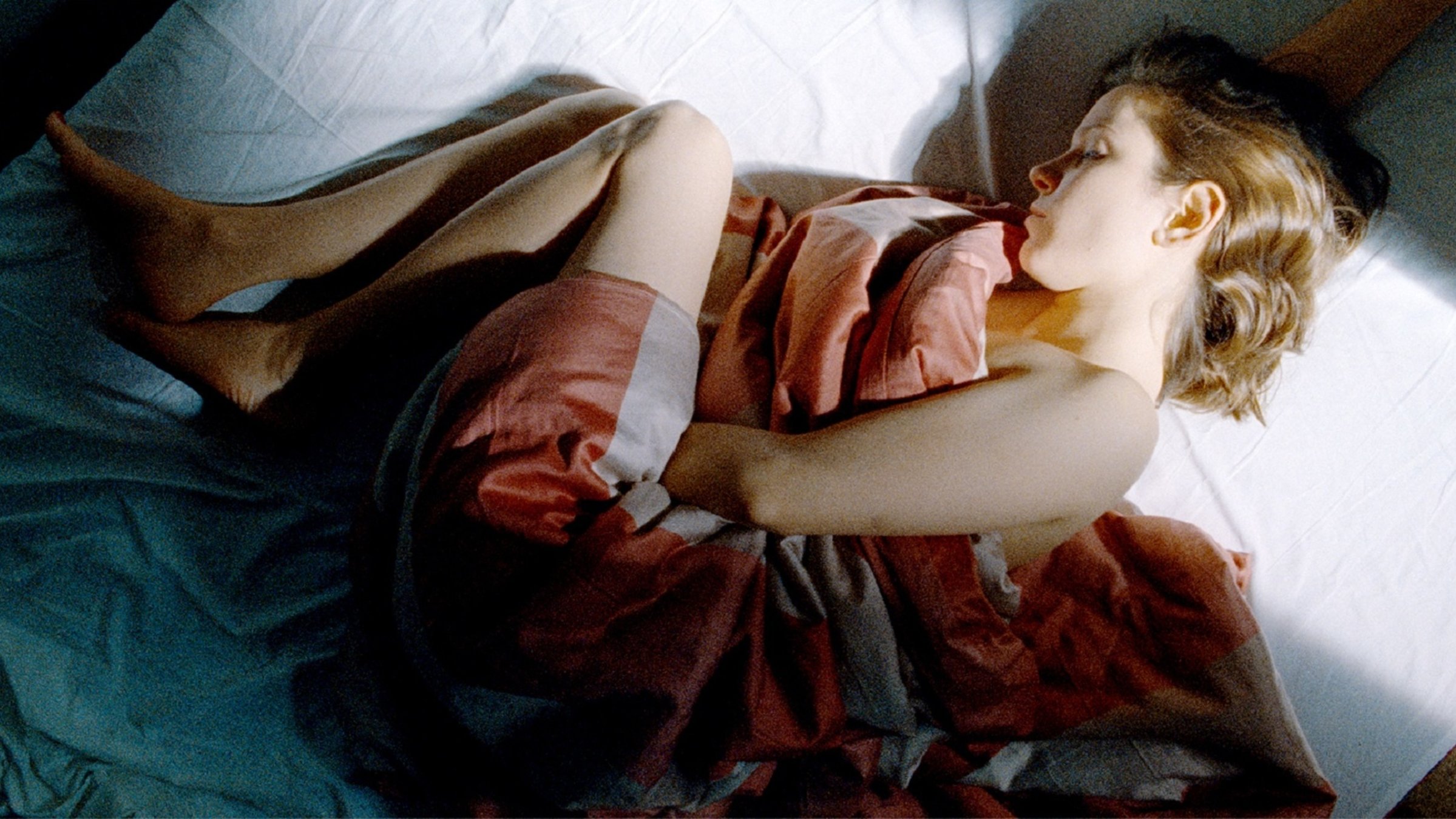 "Márta sucht János": Márta (Natasa Stork) liegt, spärlich oder gar nicht bekleidet, mit angezogenen Beinen auf einem Bett. Eine Decke bedeckt teilweise ihren Körper.