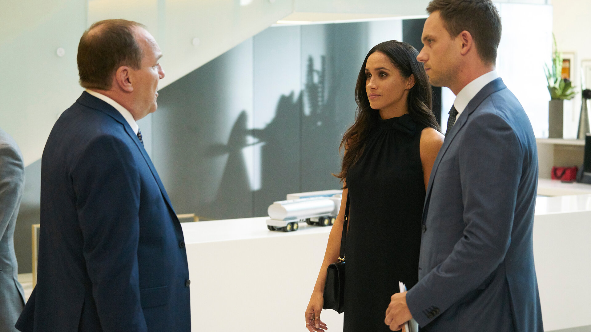 "Suits - Zerwürfnis": Die in schwarz gekleidete Rachel (Meghan Markle) und Mike (Patrick J. Adams), der in seinen Händen ein Dokument hält,  hören konzentriert Larry Burnham (Larry Day) zu, der ihnen gegenübersteht.