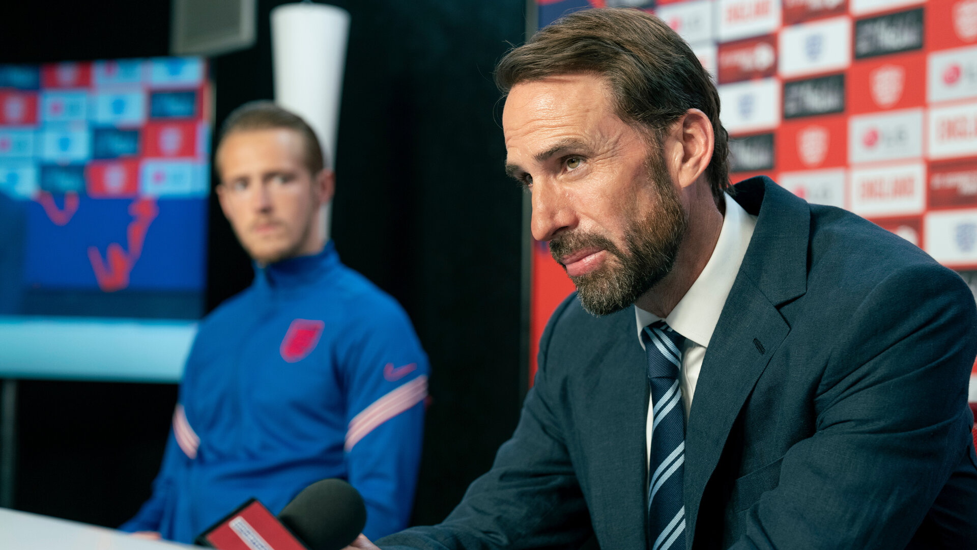 "Dear England (3)": Bei einer Pressekonferenz: Trainer Southgate (Joseph Fiennes) sitzt mit nachdenklichem Gesichtsausdruck in Anzug und Krawatte vor einem Mikrofon. Seitlich hinter ihm sitzt Will Antenbring (Harry Kane) im Trikot der englischen Nationalmannschaft.