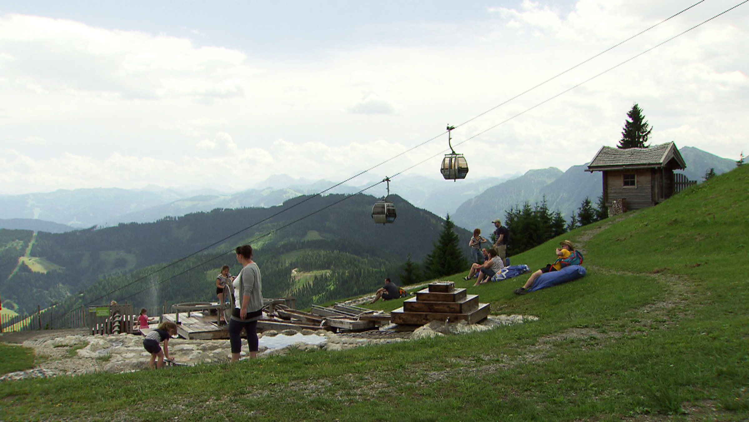 "Der Sonne entgegen Urlaub - auf Salzburgs Bergen": Mehrere Personen sitzen oder stehen auf einem Hang, dahinter läuft die Bergbahn.