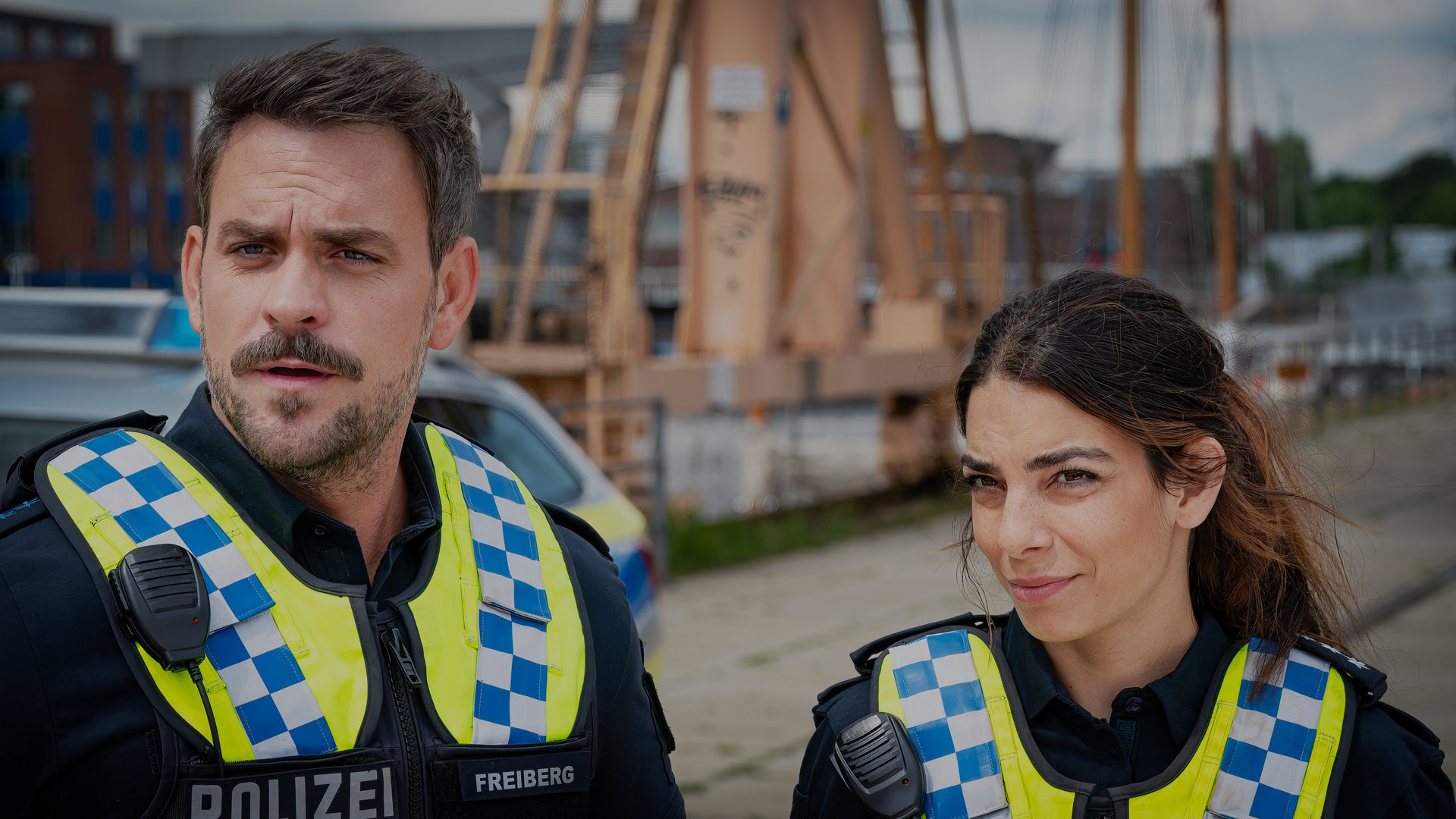 "Notruf Hafenkante - Die Neue": Großaufnahme des Polizisten Kris Freiberg (Marc Barthel) und seiner Kollegin Isa Nowak (Lilli Hollunder). Sie stehen nebeneinander und blicken auf etwas vor ihnen. Im Hintergrund liegt verschwommen die Hafenanlage.