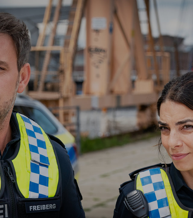 "Notruf Hafenkante - Die Neue": Großaufnahme des Polizisten Kris Freiberg (Marc Barthel) und seiner Kollegin Isa Nowak (Lilli Hollunder). Sie stehen nebeneinander und blicken auf etwas vor ihnen. Im Hintergrund liegt verschwommen die Hafenanlage.