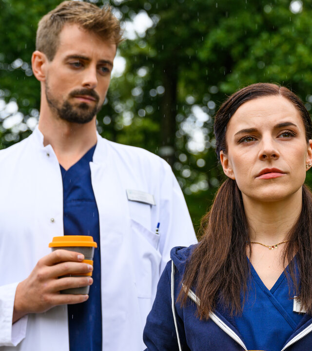 "Bettys Diagnose - Harte Landung": Im Klinikpark: Dr. Wiesner (Pablo Konrad) steht hinter Sofia (Kiara Brunken) und schaut sie an. In seiner rechten Hand hält er einen Kaffeebecher. Im Hintergrund sind Bäume zu sehen.