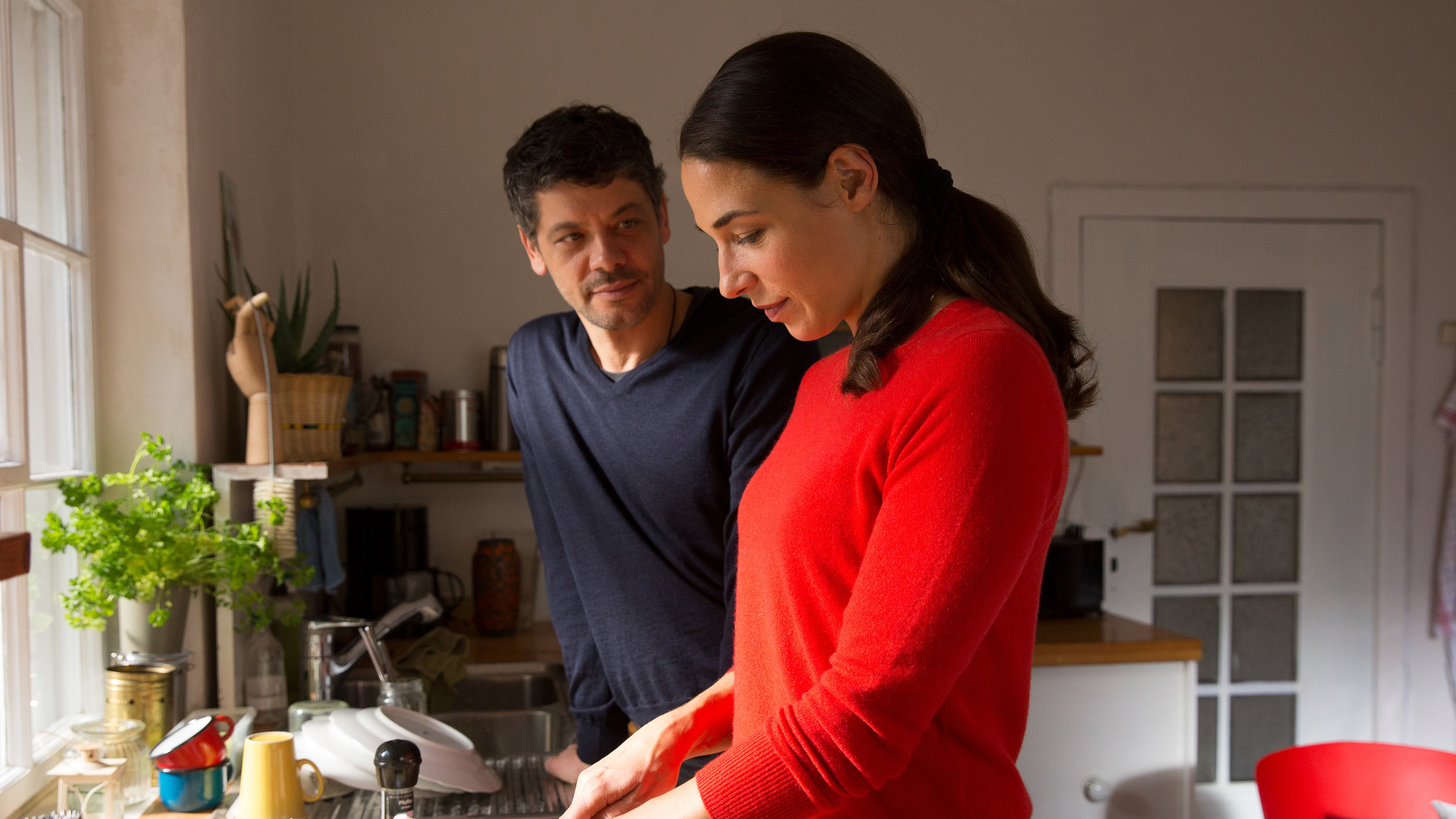 "SAFE - Episode 2": Katinka (Judith Bohle) und Tom (Carlo Ljubek) stehen nebeneinander vor einer Küchenzeile und reden. Katinka schneidet Gemüse. Auf der Küchenzeile sind diverse Tassen, Teller und andere Küchenutensilien zu sehen. Im Hintergrund sind eine Tür und ein Tisch zu sehen.