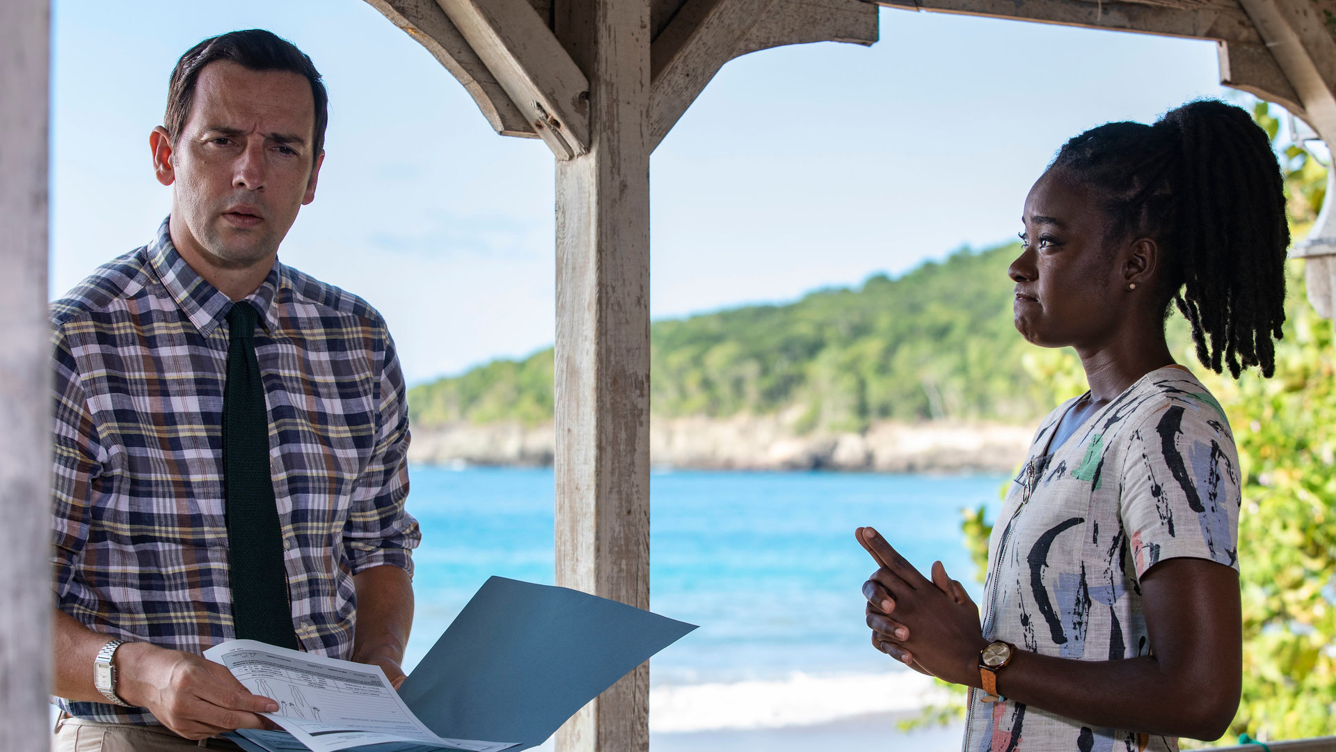 "Death in Paradise - Spiel der Könige": Naomi (Shantol Jackson) und DI Parker (Ralf Little) stehen zusammen auf einer Veranda. Während Naomi DI Parker anschaut, blickt dieser nachdenklich in die Ferne, während er eine Akte in den Händen hält.