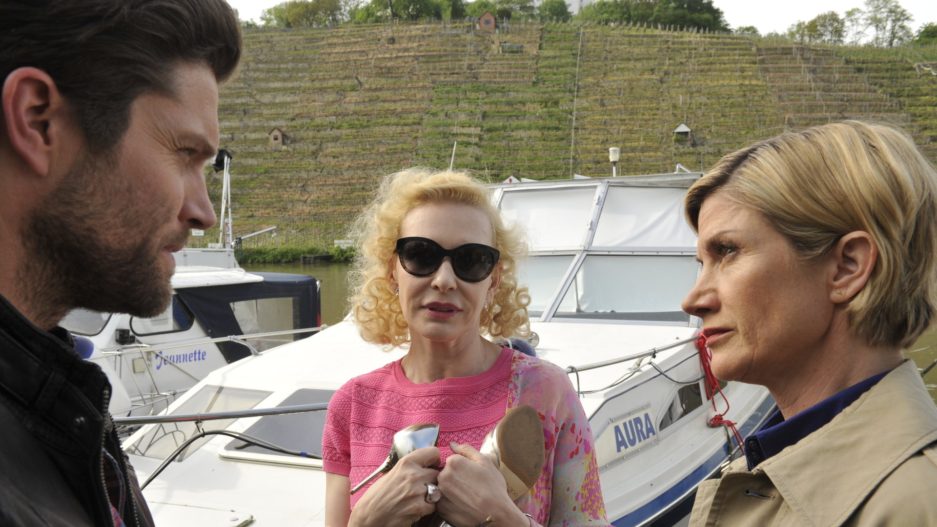 "SOKO Stuttgart - Mord mit Stil": Jo Stoll (Peter Ketnath), die Schriftstellerin Vicky Heller (Sunnyi Melles) und Martina Seiffert (Astrid M. Fünderich) stehen am Neckar vor der kleinen weißen Yacht "Aura". Vicky hält ihre High Heels in den Händen an die Brust gedrückt.
