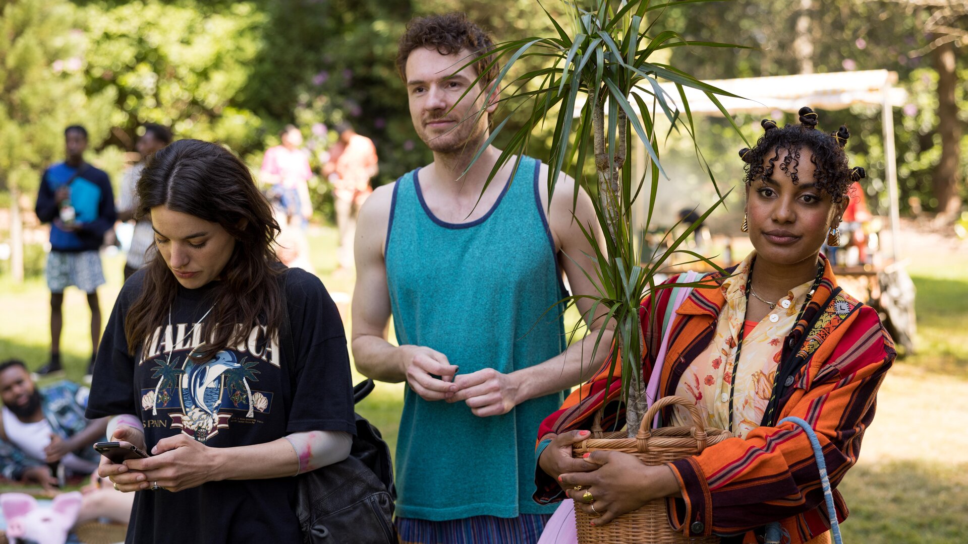 This Is Gonna Be Great - Höhepunkt": Hanna (Mona Vojacek Koper), David (Rein Mulder) und Aisha (Abigail Abraham) stehen nebeneinander im Grünen. Im Hintergrund befinden sich weitere Menschen. Hanna hantiert mit ihrem Handy. Aisha hält eine große Topfpflanze im Arm.