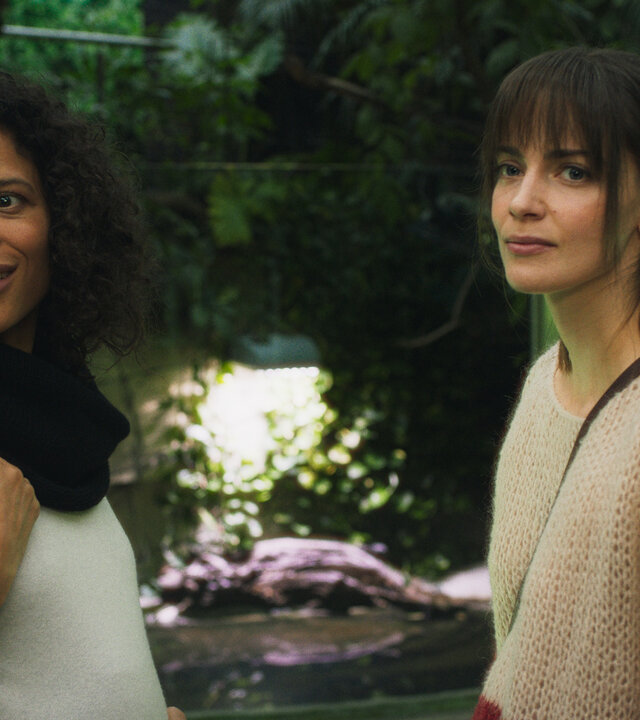 "Bauchgefühl - Zoe": Zoe (Davina Donaldson) und Lena (Laura Berlin) stehen im Zoo vor einem Terrarium und gucken gemeinsam auf etwas in der Ferne. Beide tragen Taschen über den Schultern. Zoe wirkt positiv, aber Lena verschlossen.