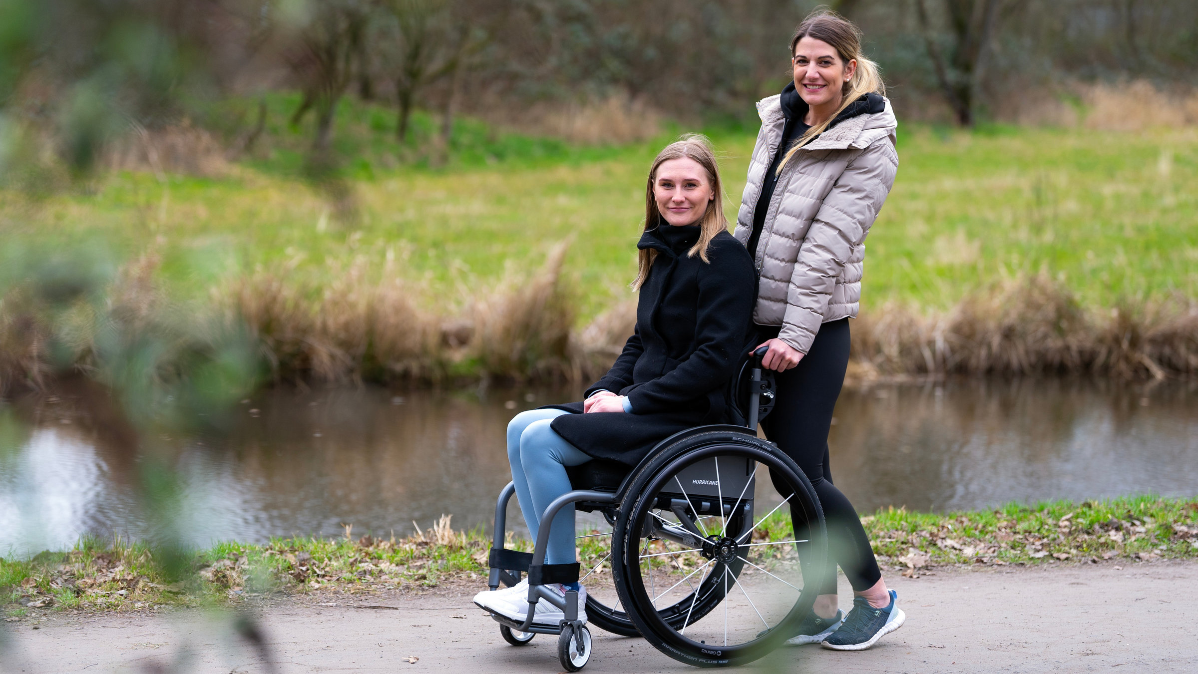 "einfach Mensch: Sina und Tina - eine besondere Freundschaft": Tina im Rollstuhl und Sina zusammen auf einem Gehweg an einem Fluss.