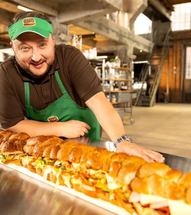 "besseresser: Die Tricks von McDonald´s und Co.": Produktentwickler Sebastian Lege) trägt eine grüne Schürze und eine grüne Schirmmütze und steht neben einem meterlangen belegten Sandwich.