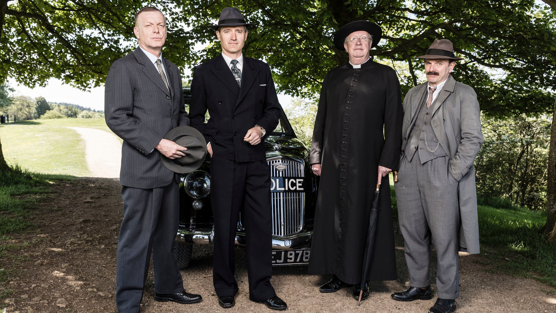 "Father Brown - Der Turm der verlorenen Seelen": Inspector Valentine (Hugo Speer), Chief Inspector Sullivan (Tom Chambers), Father Brown (Mark Williams) und Inspector Mallory (Jack Deam) stehen vor dem Polizeiwagen.