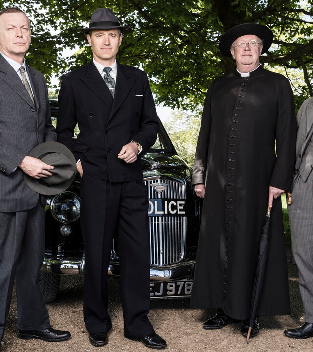 "Father Brown - Der Turm der verlorenen Seelen": Inspector Valentine (Hugo Speer), Chief Inspector Sullivan (Tom Chambers), Father Brown (Mark Williams) und Inspector Mallory (Jack Deam) stehen vor dem Polizeiwagen.