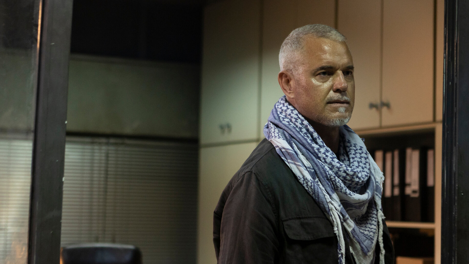 "Kabul - Flucht aus der Hölle: Ungewöhnliche Weggefährten": Martin (Eric Dane) steht im Rahmen der Glastür zu einem Büro und schaut ernst zur Seite. Er trägt ein typisches afghanisches Tuch um den Hals.