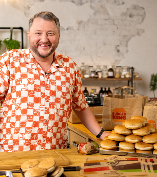"besseresser: Die Wahrheit über McDonald's & Co. - Sebastian Lege deckt auf": Sebastian Lege steht in seiner Küche vor einer Arbeitsfläche. Vor ihm liegen Burger Brötchen und Käsescheiben sowie Verpackungspapier. Neben ihm stapeln weitere Burger Brötchen. Im Hintergrund stehen Burger King Tüten.