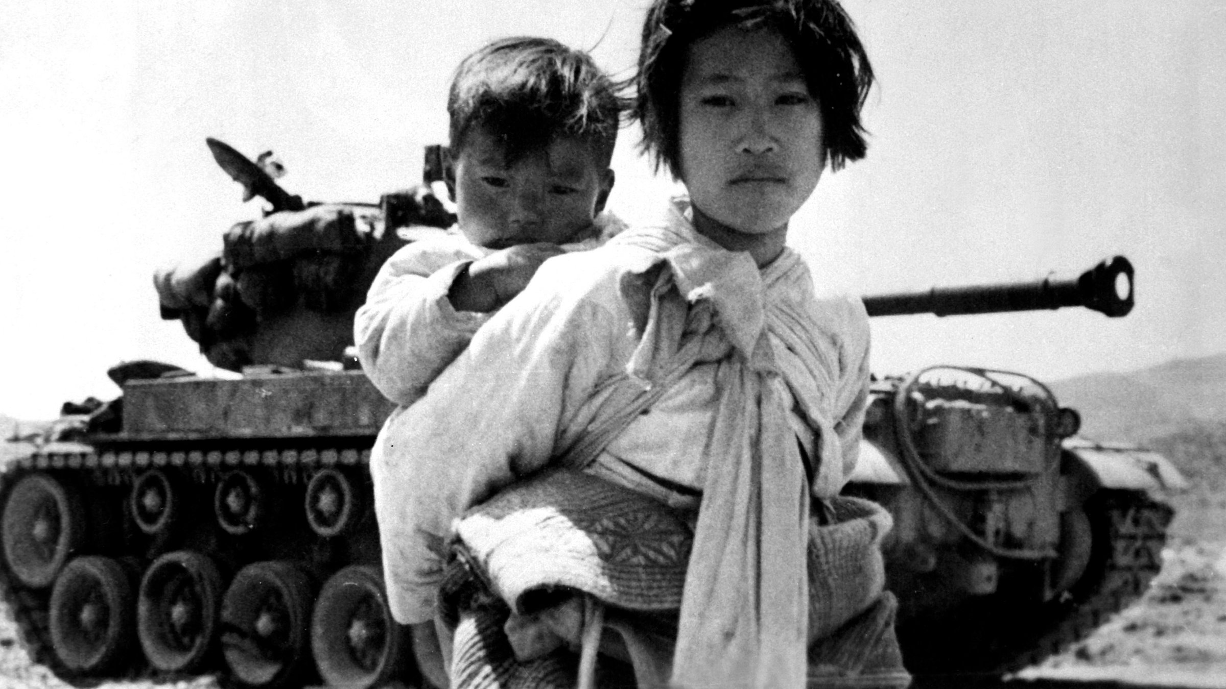 "Pulverfass Korea - Konflikt ohne Ende": Schwarz-Weiß-Foto: Ein junges Mädchen mit einem Kleinkind auf den Rücken geschnallt steht vor einem Panzer.