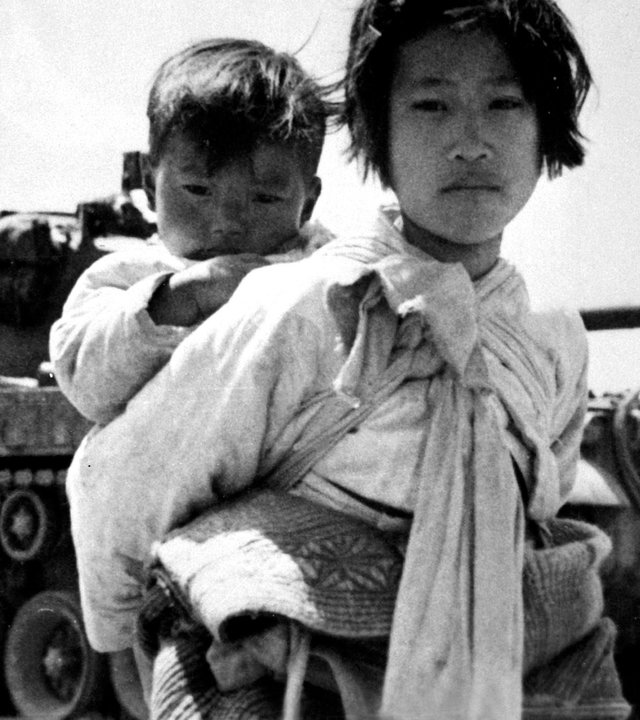"Pulverfass Korea - Konflikt ohne Ende": Schwarz-Weiß-Foto: Ein junges Mädchen mit einem Kleinkind auf den Rücken geschnallt steht vor einem Panzer.