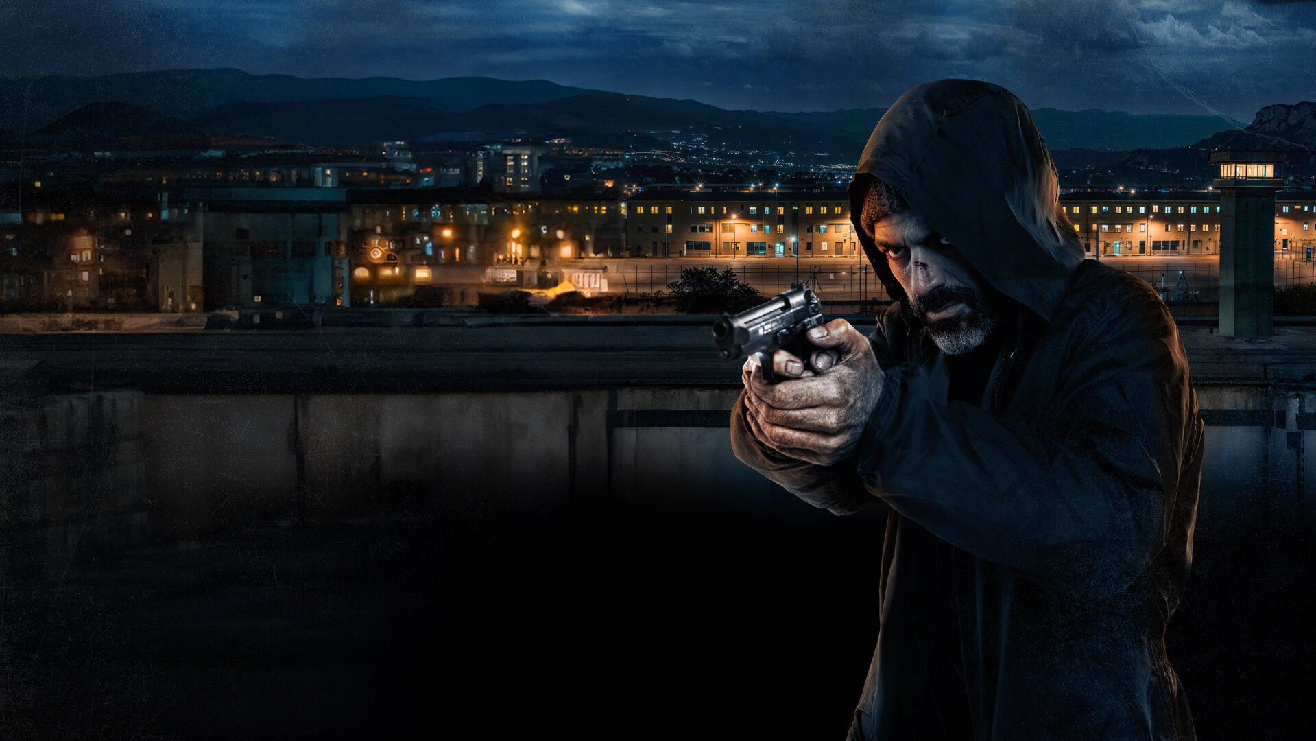 "Verschwunden auf Sardinien": Sante Moras (Luca Argentero) zielt mit gezogener Pistole schräg in Richtung Kamera. Er trägt einen dunklen Kapuzenpullover. Im Hintergrund ist die Totale eines Gefängnisses in der Dunkelheit zu sehen, die Fenster sind hell erleuchtet. Zwischen dem Gebäudekomplex und Sante befindet sich eine Mauer.