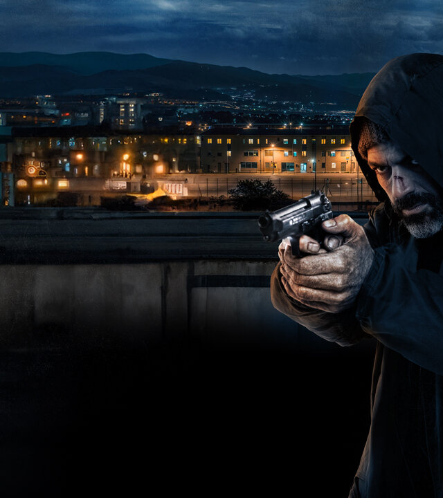 "Verschwunden auf Sardinien": Sante Moras (Luca Argentero) zielt mit gezogener Pistole schräg in Richtung Kamera. Er trägt einen dunklen Kapuzenpullover. Im Hintergrund ist die Totale eines Gefängnisses in der Dunkelheit zu sehen, die Fenster sind hell erleuchtet. Zwischen dem Gebäudekomplex und Sante befindet sich eine Mauer.