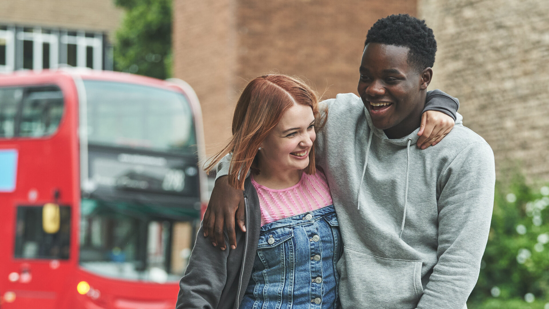 "Boarders - Welcome to St. Gilbert's - Brownies & Betrayal": Beatrix (Tallulah Greive) und Jaheim (Josh Tedeku) gehen Arm in Arm einer Straße entlang. Beide lachen. Im Hintergrund steht ein roter Bus.