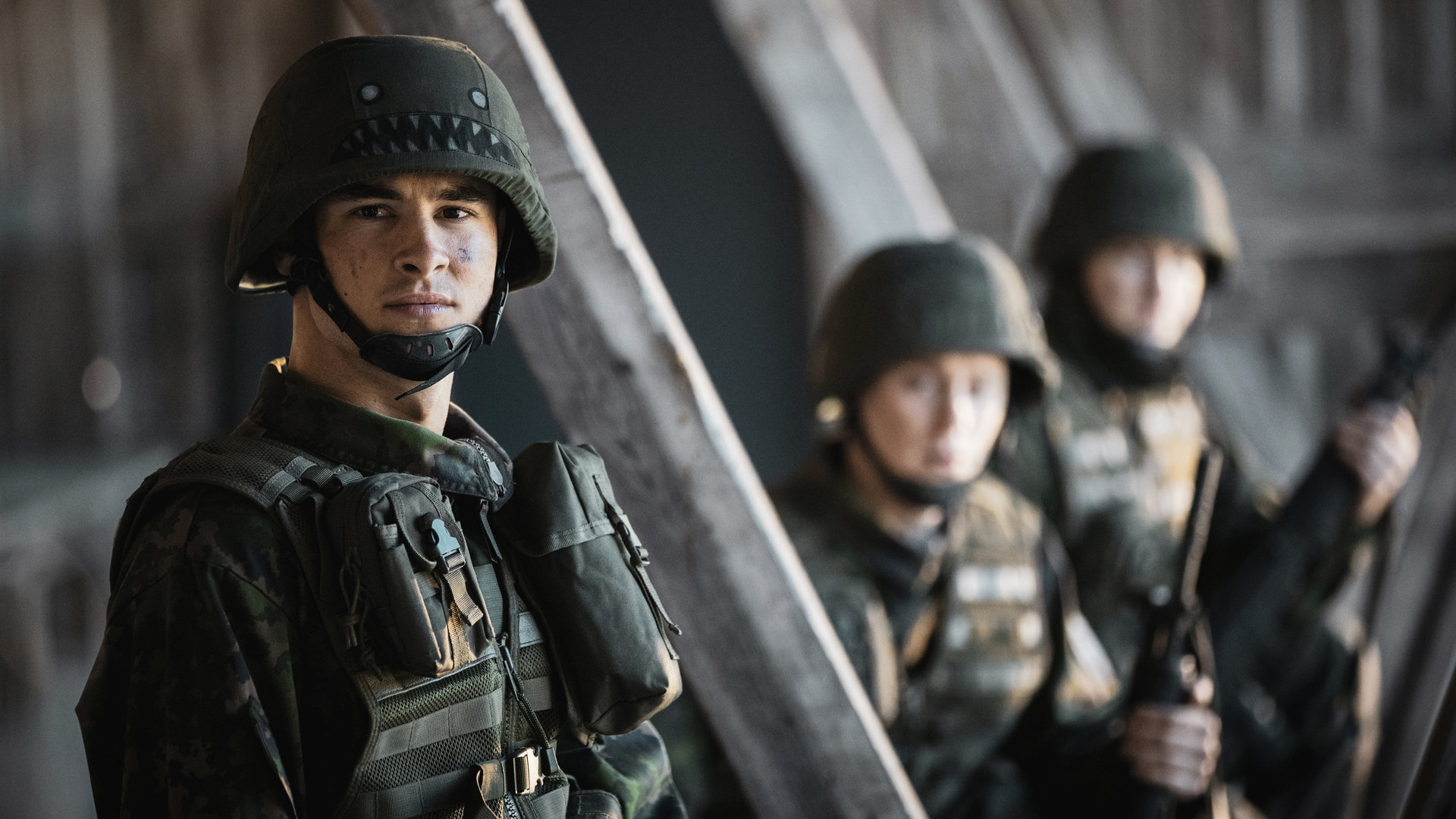 "Californian Commando - Entführt": Van (Kian Lawley) steht in militärischer Tarnuniform neben einem Holzpfahl und zwei Kameraden und schaut konzentriert zur Seite.