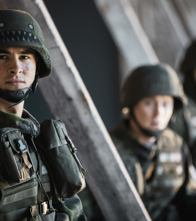 "Californian Commando - Entführt": Van (Kian Lawley) steht in militärischer Tarnuniform neben einem Holzpfahl und zwei Kameraden und schaut konzentriert zur Seite.