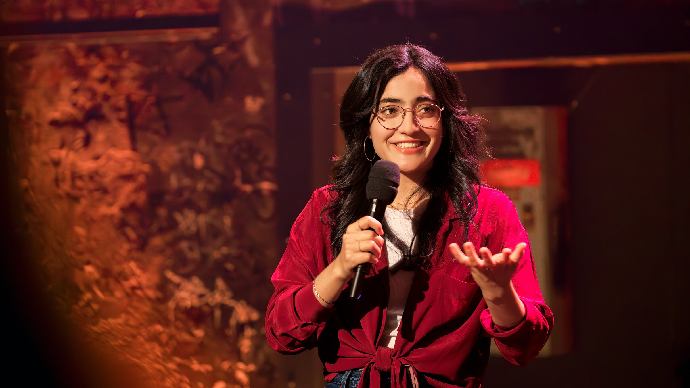 "Till Reiners´ Happy Hour - Takeover - Primetime-Comedy mit Ana Lucía und Gästen": Die Stand-Up-Comedienne Ana Lucía steht mit einem Mikrofon in der Hand auf der Bühne und spricht zum Publikum