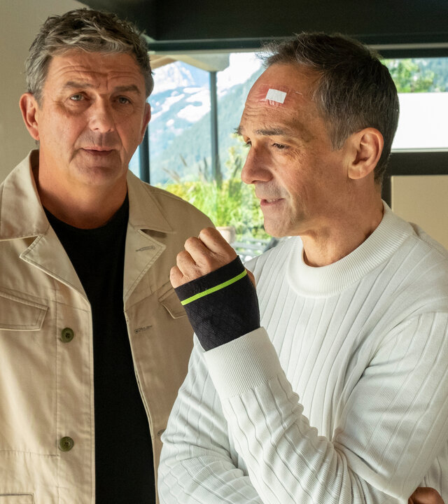 "Der Bergdoktor: Unverzeihlich": Im Haus von Gero Jung: Nachdenklich, beide Händen in den Hosentaschen, steht Dr. Martin Gruber (Hans Sigl) neben Gero Jung (Harry Blank). Dieser blickt entschlossen aus dem Fenster. An seiner Stirn klebt ein Pflaster und an seiner Hand trägt er eine Manschette.