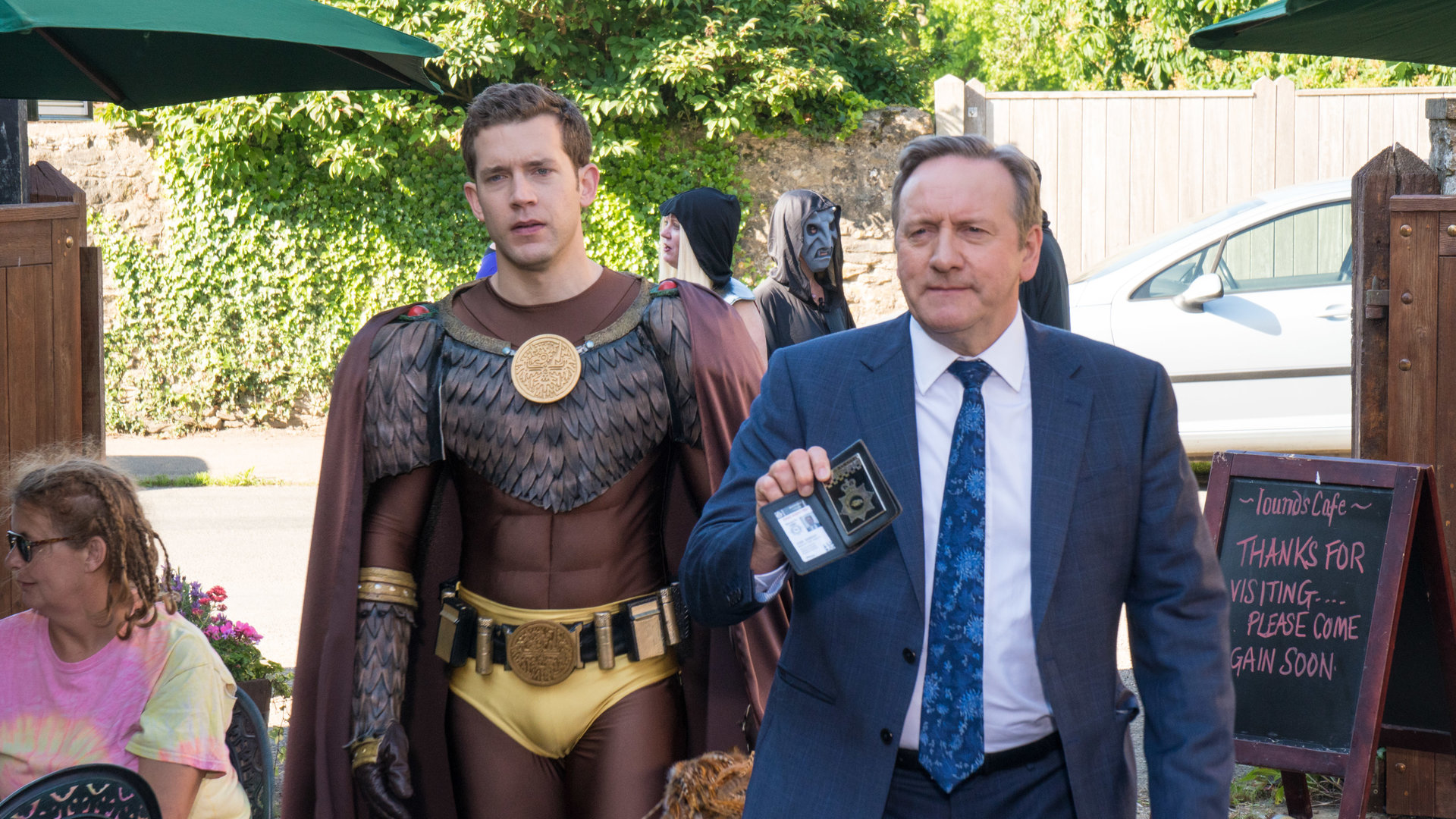 "Inspector Barnaby - Was geschah wirklich auf Schloss Argo?": DCI John Barnaby (Neil Dudgeon), geschmackvoll gekleidet in Anzug und Krawatte, geht auf die Terrasse eines Cafés und zeigt seinen Dienstausweis vor. Neben ihm geht sein Assistent Jamie Winter (Nick Hendrix), gekleidet in einem bronzefarbenen Superhelden-Kostüm mit Umhang und federartigen Applikationen.