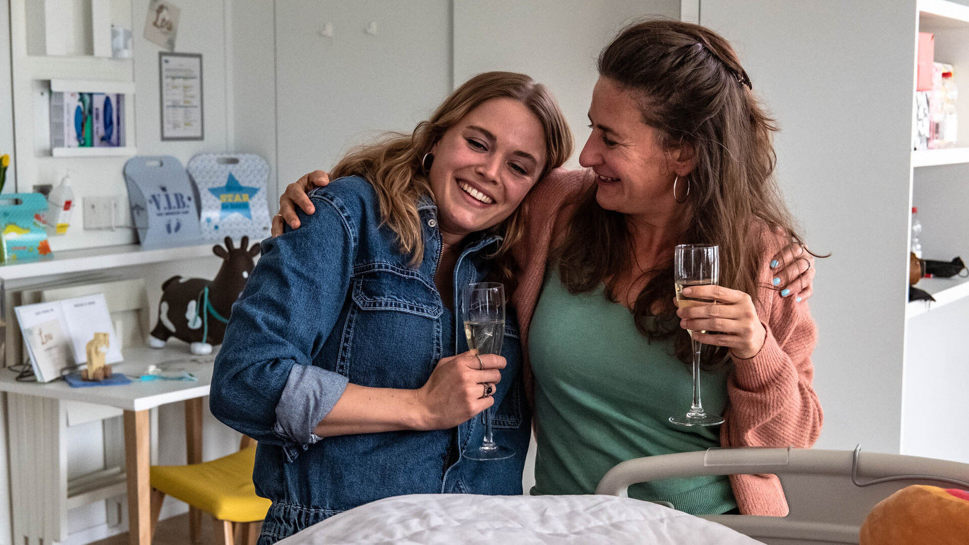 "The Club - Der Club, zu dem du gehörst": Charlie (Roos Dickmann) und Kirstie (Janne Desmet) stehen lachend mit einem Sektglas in der Hand vor einem Krankenhausbett und umarmen sich dabei.