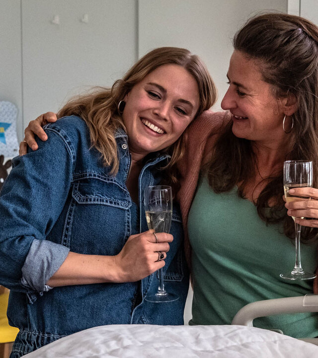 "The Club - Der Club, zu dem du gehörst": Charlie (Roos Dickmann) und Kirstie (Janne Desmet) stehen lachend mit einem Sektglas in der Hand vor einem Krankenhausbett und umarmen sich dabei.