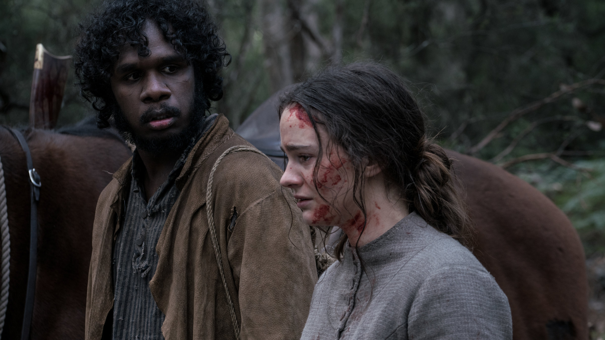 "The Nightingale": Clare (Aisling Franciosi) steht mit Blut verschmiertem Gesicht neben ihrem Begleiter, dem Aborigine Billy (Baykali Ganambarr), der sie entgeistert anschaut. Im Hintergrund ist der Rücken eines Pferdes im Wald zu sehen.