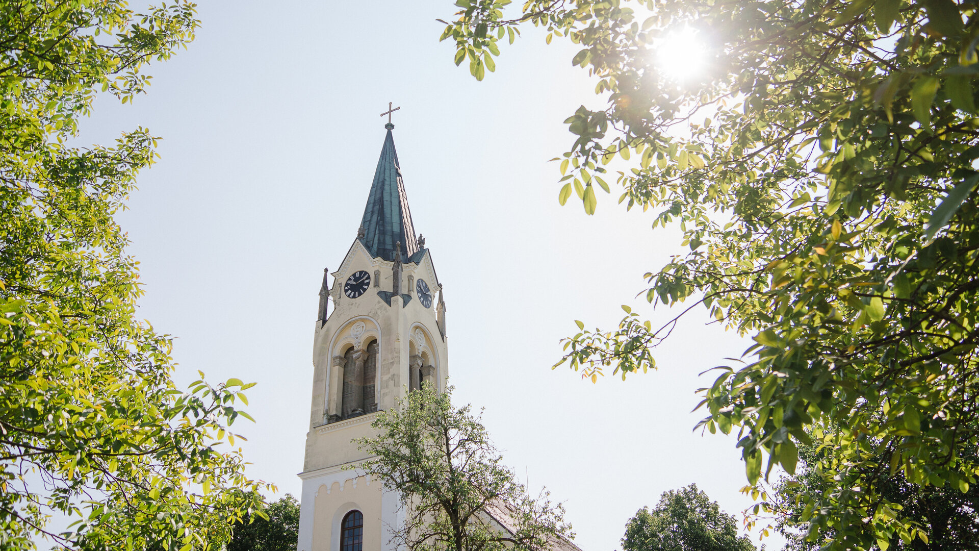 "Evangelischer Gottesdienst zum Erntedankfest - Kraut und Rüben": Untersicht: Zwischen den Laubkronen der Bäume ragt der weiße Turm der "Kirche im Feld" bei Sonnenschein in den Himmel. Die Kirche wurde im neuromanischen Stil erbaut.