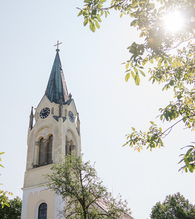 "Evangelischer Gottesdienst zum Erntedankfest - Kraut und Rüben": Untersicht: Zwischen den Laubkronen der Bäume ragt der weiße Turm der "Kirche im Feld" bei Sonnenschein in den Himmel. Die Kirche wurde im neuromanischen Stil erbaut.