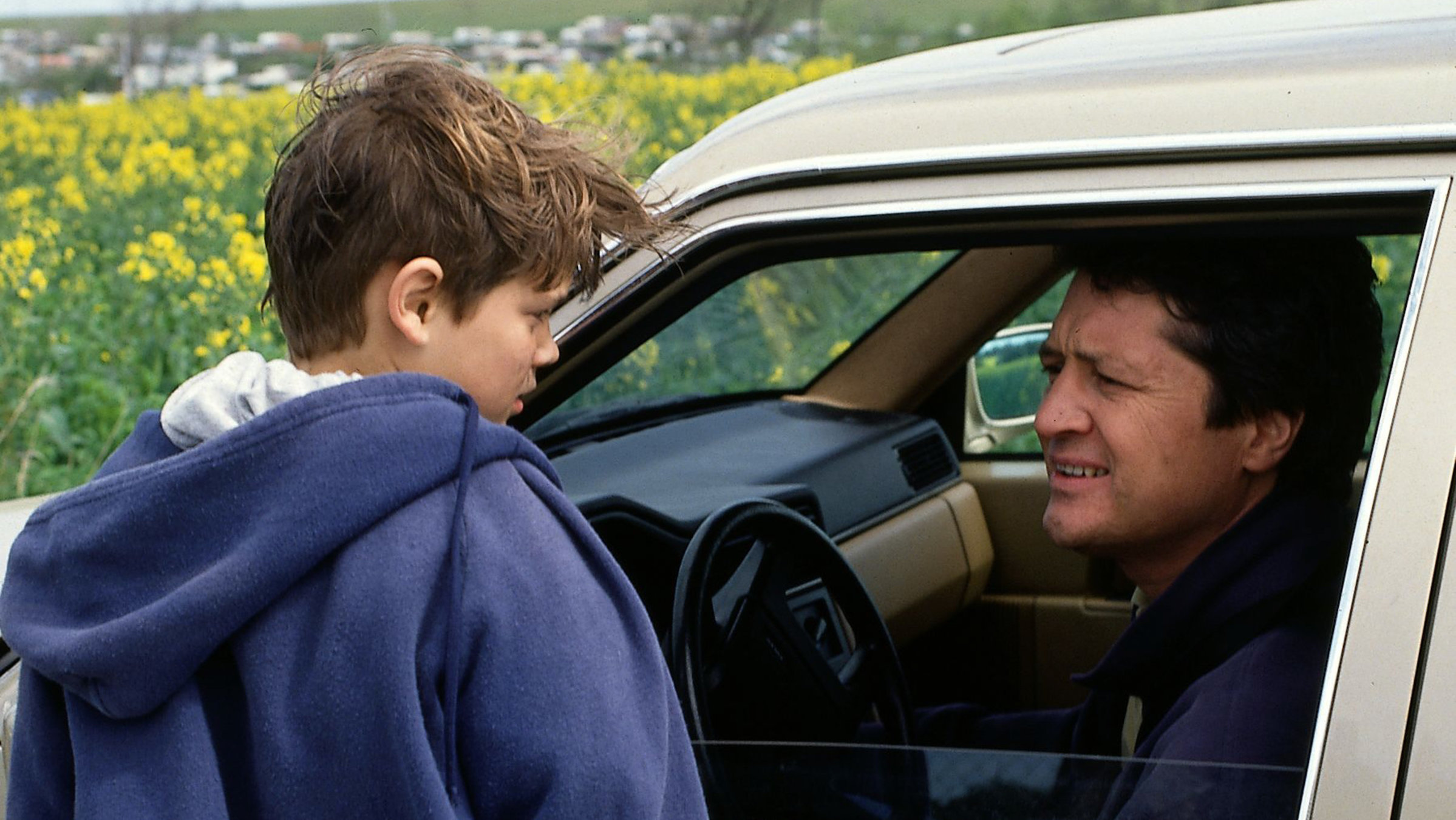 "Der Landarzt - Der Tod des Dr.  Mattiesen": Dr. Mattiesen (Christian Quadflieg) sitzt in seinem stehenden Auto. Er hat die Fensterscheibe offen und spricht mit einem Jungen (Antonio Wannek). Beide schauen besorgt. Im Hintergrund eine Wiese mit gelben Rapsblüten.
