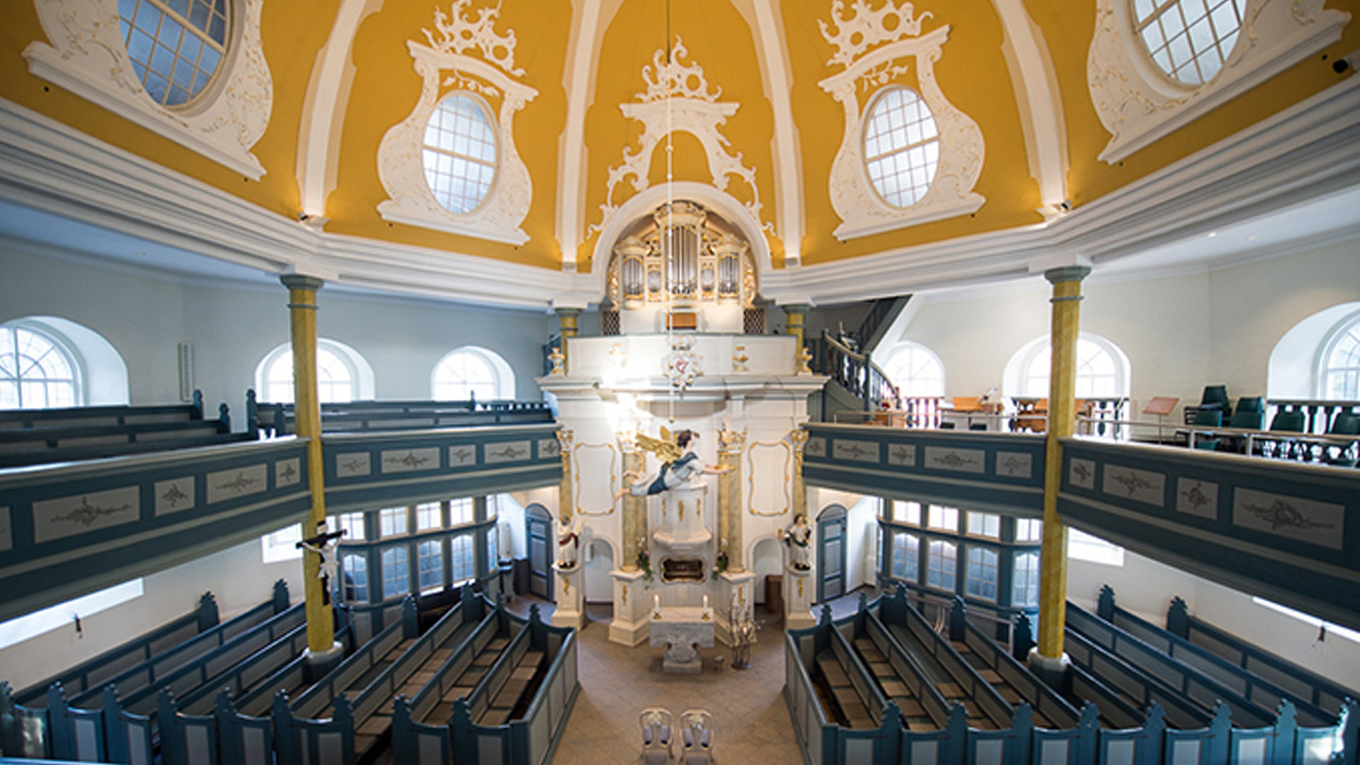 "Evangelischer Gottesdienst zur Christnacht - In neuem Licht": Blick in den Innenraum der spätbarocken Kirche am Markt in Hamburg-Niendorf: Ein hölzerner Engel schwebt hoch über dem achteckigen Altarraum. Das kuppelartige, ockerfarbene Mansardendach wird von weiß umrandeten Fenstern durchbrochen. Das Gestühl und Balkongeländer ist in Grau-Blautönen gehalten.