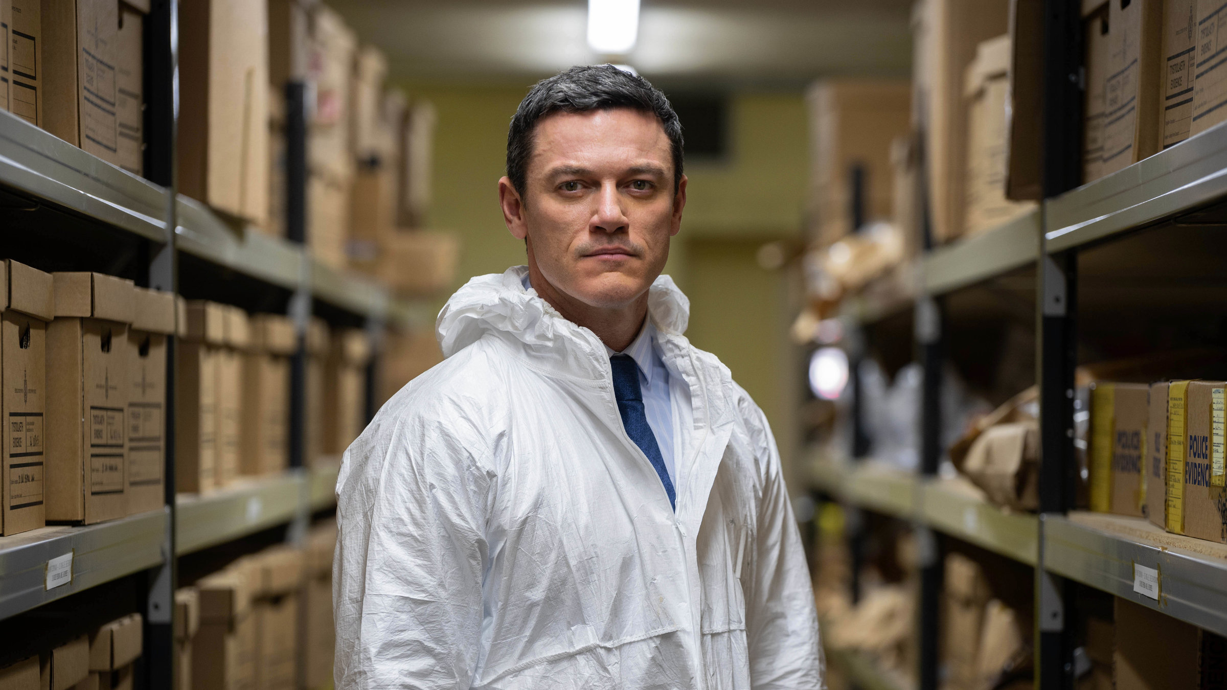 "Die Pembrokeshire Morde (1)": DCS Steve Wilkins (Luke Evans) steht, mit einem weißen Schutzanzug bekleidet, in der Asservatenkammer und sieht in die Kamera.
