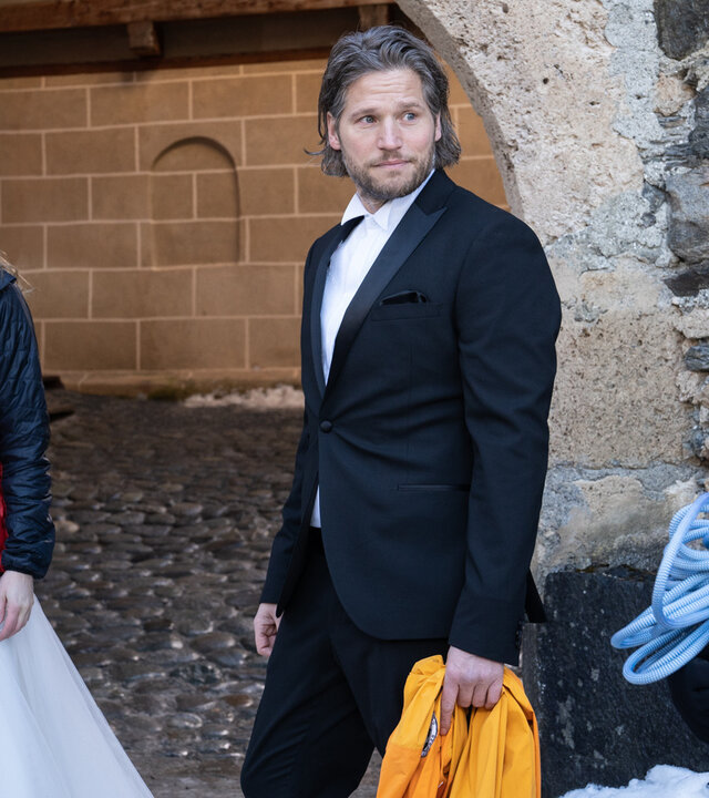 "Die Bergretter: Dieses eine Leben": Unter einem Torbogen steht Katharina (Luise Bähr) in einem weißen Brautkleid, darüber ihre Rettungsjacke und über die Schulter tragend ihren Arztkoffer. Neben ihr steht Markus (Sebastian Ströbel) in einem schwarzen Anzug. Beide blicken zu Bergretter Rudi (Michael Pascher).