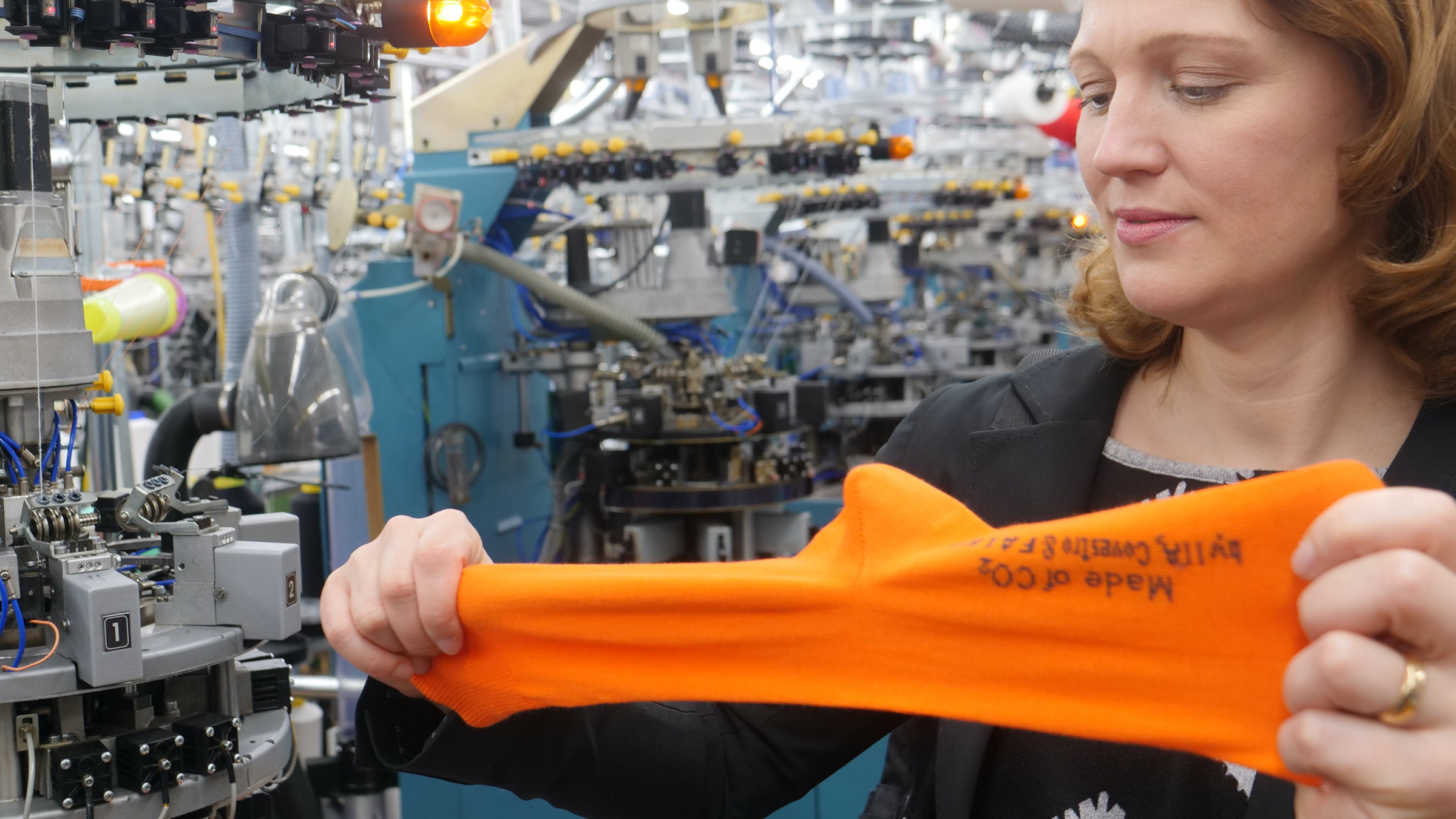 "plan b: Socken aus CO2 - Wie ein Klimakiller nützlich werden kann": Eine Frau steht neben Maschinen und zieht an einer orangefarbenen Socke.