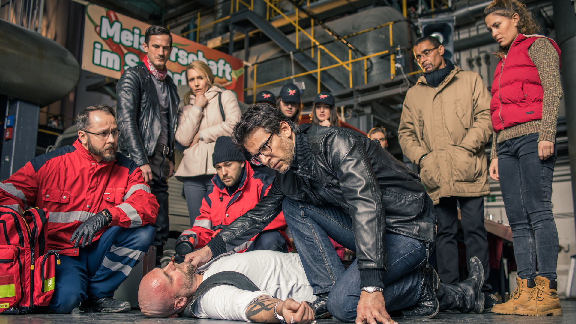"SOKO München - Schärfer als die Polizei erlaubt": Auf einer Contest-Veranstaltung in einer Halle liegt eine männliche Leiche auf dem Boden, Dr. Weissenböck (Florian Odendahl) sitz daneben in der Hocke und prüft den Puls am Hals, gegenüber sitzen zwei Sanitäter in der Hocke. Im Hintergrund stehen Toni Bischoff (Amanda da Gloria), Franz Ainfachnur (Christofer v. Beau) und Komparsen.