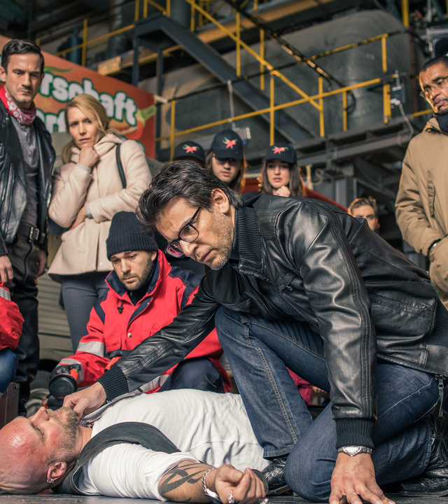 "SOKO München - Schärfer als die Polizei erlaubt": Auf einer Contest-Veranstaltung in einer Halle liegt eine männliche Leiche auf dem Boden, Dr. Weissenböck (Florian Odendahl) sitz daneben in der Hocke und prüft den Puls am Hals, gegenüber sitzen zwei Sanitäter in der Hocke. Im Hintergrund stehen Toni Bischoff (Amanda da Gloria), Franz Ainfachnur (Christofer v. Beau) und Komparsen.