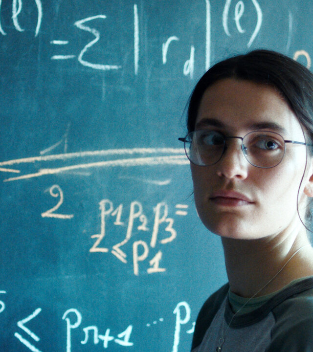"Die Gleichung ihres Lebens": Nahaufnahme: Marguerite (Ella Rumpf) steht neben einer Tafel voll mathematischer Formeln und blickt auf etwas hinter der Kamera.