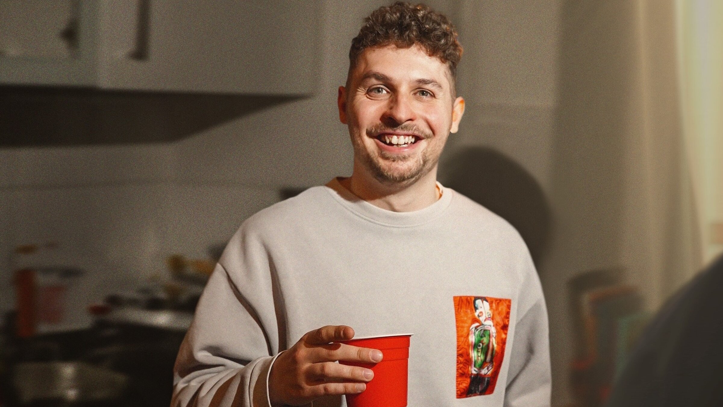 "Fabian Lampert: Einer dieser Abende": Fabian Lampert steht mit einem roten Getränkebecher aus Plastik in der Küche und schaut in die Kamera.