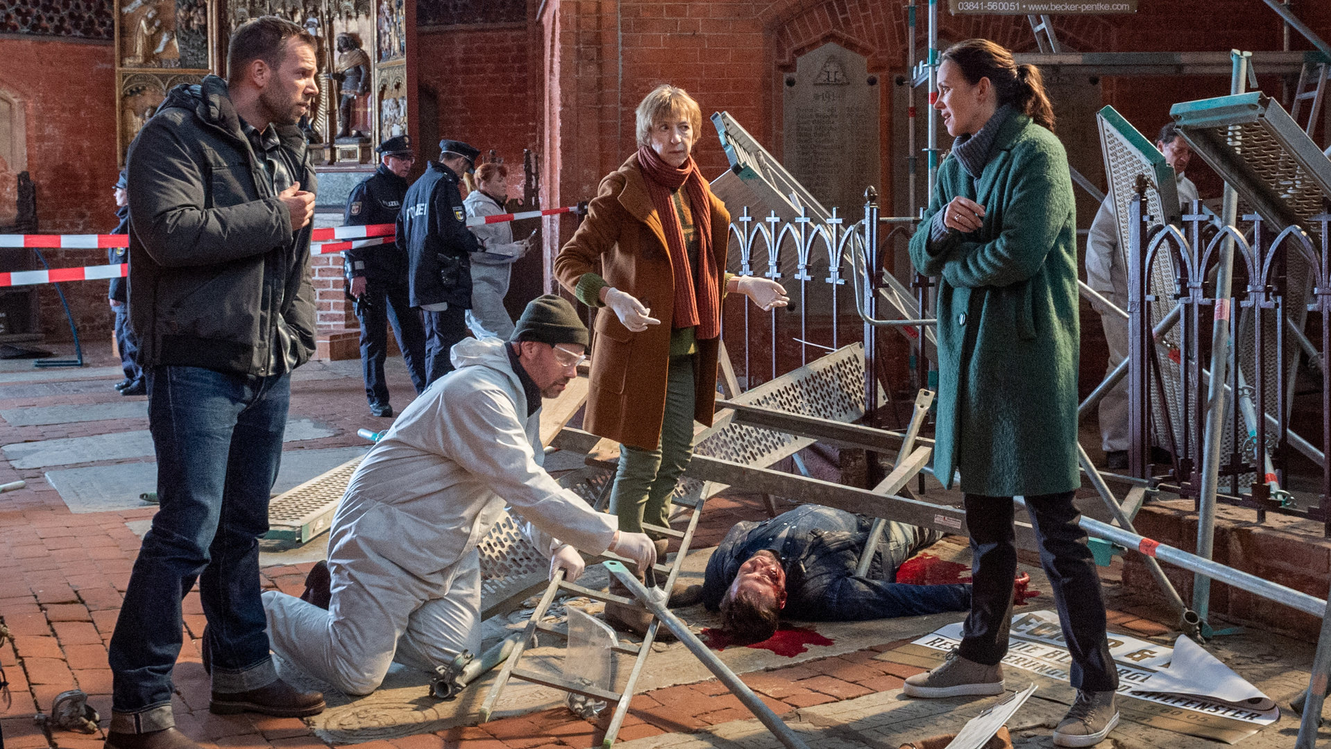 „SOKO Wismar – Bleiverglasung“: In einer Kirche liegt Jonas Pentke tot (Johannes Hlépas) unter einem umgestürzten Baugerüst. Kriminaltechniker Roland (Christian Intorp) kniet links von ihm und räumt Teile des Gerüsts beiseite. Rings um den Toten stehen Lars Pöhlmann (Dominic Boeer), Helene Sturbeck (Katharina Blaschke) und Karoline Joost (Nike Fuhrmann). Im Hintergrund erstreckt sich das Backsteingemäuer der Kirche. Weiter rechts hängt eine Jesusfigur an einem Kreuz.