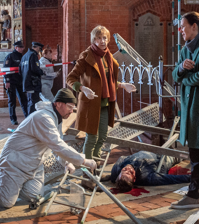 „SOKO Wismar – Bleiverglasung“: In einer Kirche liegt Jonas Pentke tot (Johannes Hlépas) unter einem umgestürzten Baugerüst. Kriminaltechniker Roland (Christian Intorp) kniet links von ihm und räumt Teile des Gerüsts beiseite. Rings um den Toten stehen Lars Pöhlmann (Dominic Boeer), Helene Sturbeck (Katharina Blaschke) und Karoline Joost (Nike Fuhrmann). Im Hintergrund erstreckt sich das Backsteingemäuer der Kirche. Weiter rechts hängt eine Jesusfigur an einem Kreuz.