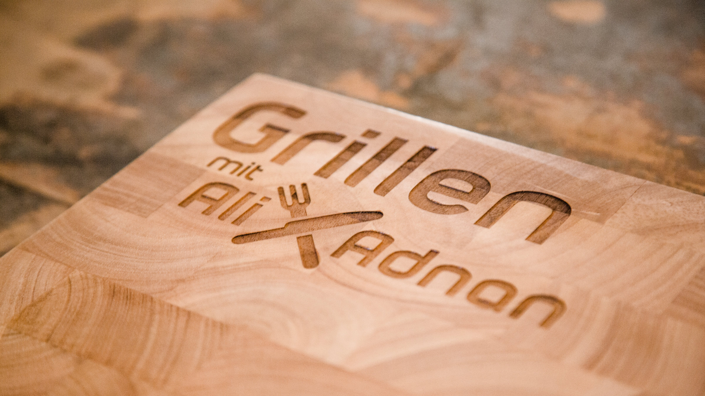 "Grillen mit Ali und Adnan": "Grillen mit Ali und Adnan"-Sendungslogo auf einem Holzbrett.