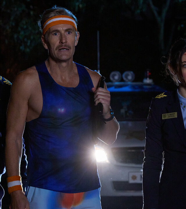 "Gnomes - Angriff der Killerzwerge - Armee der Zwerge": Nachts stehen Arnold (Asa Butterfield), Harvey (Brett Tucker) und Ellie (Megan Smart) im Scheinwerferlicht eines Polizeiwagens im Freien. Arnold blickt schockiert nach vorn. Neben ihm steht Harvey in bunter Sportkleidung mit erhobener Hand, während Ellie mit einer blutigen Platzwunde an der Stirn ungläubig in dieselbe Richtung starrt. Das Licht des Streifenwagens beleuchtet die drei von hinten.