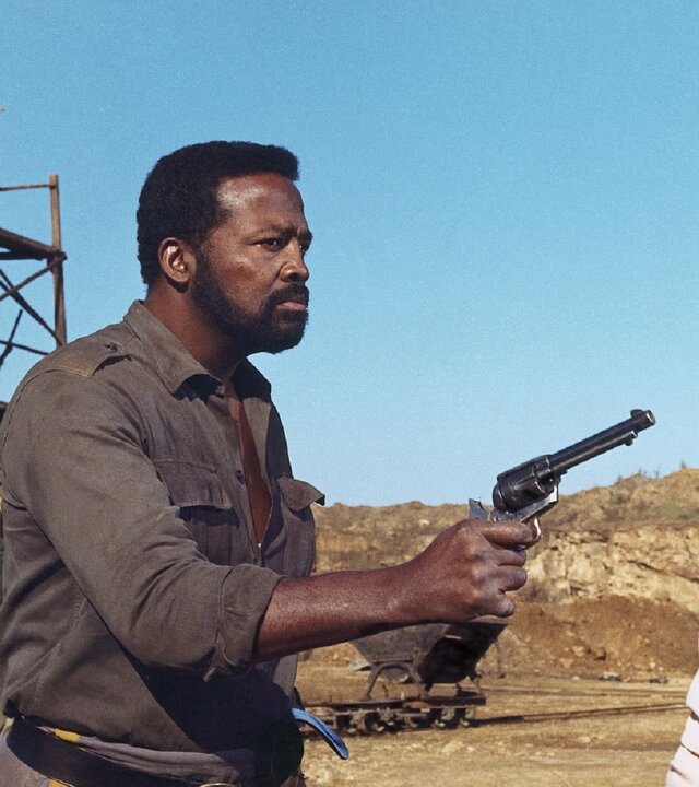 "Die letzte Rechnung zahlst du selbst": In einer wüstenartigen Landschaft hält Al (Al Hoosman) eine Pistole auf Billy (Lee Van Cleef) gerichtet, der ihm mit ernster Miene gegenübersteht, während Ben (Antonio Sabàto) im Hintergrund mit einem Gewehr im Anschlag bereitsteht. Hinter ihnen befinden sich ein hölzernes Baugerüst und ein Pferdewagen.