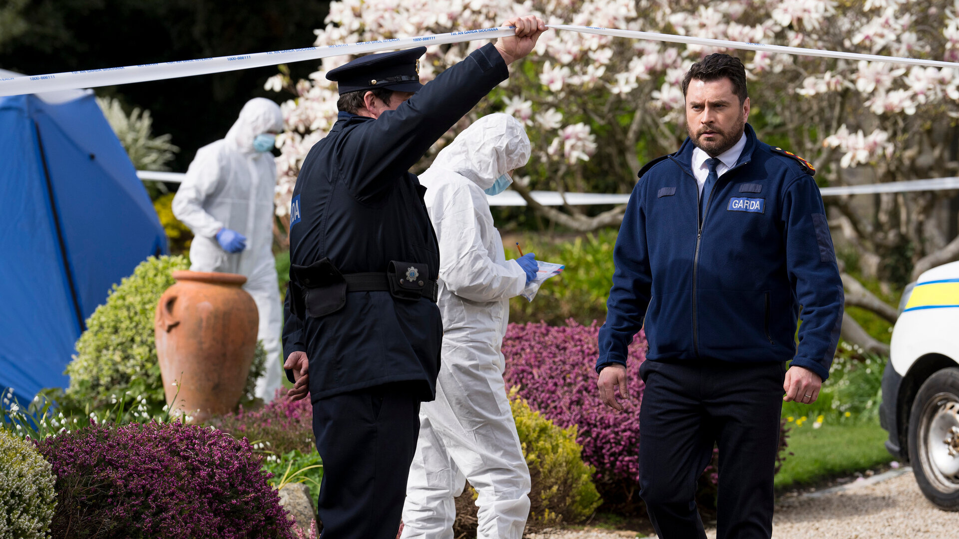 "Harry Wild - Mörderjagd in Dublin - Unser Dorf soll schöner morden": Charlie Wild (Kevin Ryan) ist auf dem Weg zum Tatort. Er läuft unter einem weißen Absperrband hindurch, welches ein seitlich stehender Polizist für ihn hochhält. Im Hintergrund arbeiten zwei Mitglieder der Spurensicherung. Sie sind mit weißen Papieranzügen, Mundschutz, blauen Einweghandschuhen und blauen Einwegschuhüberziehern bekleidet. Dahinter steht ein blaues Zelt in einem blühenden Garten.