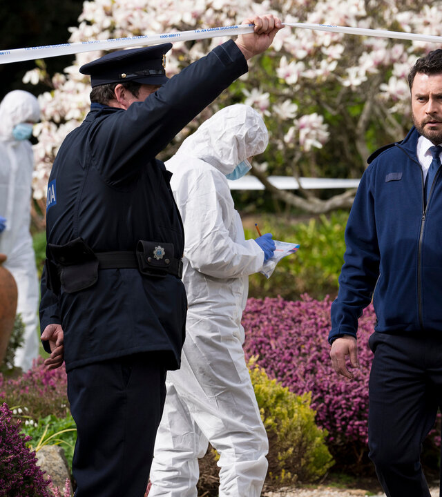 "Harry Wild - Mörderjagd in Dublin - Unser Dorf soll schöner morden": Charlie Wild (Kevin Ryan) ist auf dem Weg zum Tatort. Er läuft unter einem weißen Absperrband hindurch, welches ein seitlich stehender Polizist für ihn hochhält. Im Hintergrund arbeiten zwei Mitglieder der Spurensicherung. Sie sind mit weißen Papieranzügen, Mundschutz, blauen Einweghandschuhen und blauen Einwegschuhüberziehern bekleidet. Dahinter steht ein blaues Zelt in einem blühenden Garten.
